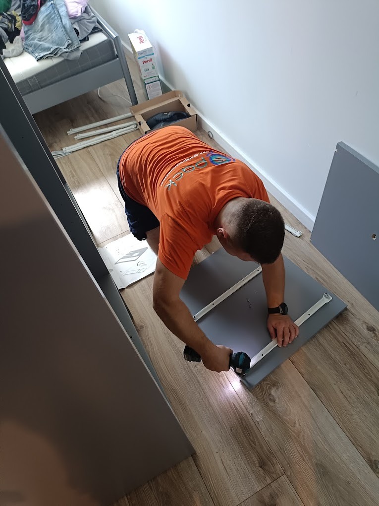 Monter skręca mebel na podłodze z paneli, używając wkrętarki akumulatorowej, widoczny fragment łóżka piętrowego i opakowanie po proszku do prania.