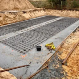 Przygotowanie zbrojenia na betonowej płycie fundamentowej, dwóch pracowników w odblaskowych kamizelkach przy pracy, widoczny szalunek z desek, wiadro, miara zwijana i młotek.