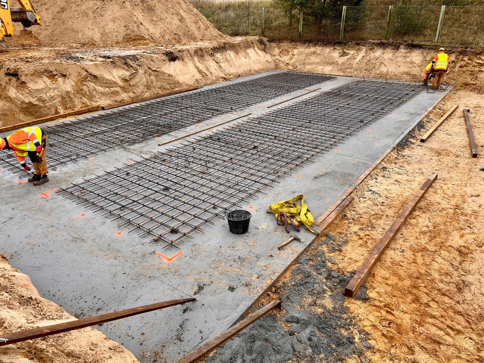 Przygotowanie zbrojenia na betonowej płycie fundamentowej, dwóch pracowników w odblaskowych kamizelkach przy pracy, widoczny szalunek z desek, wiadro, miara zwijana i młotek.