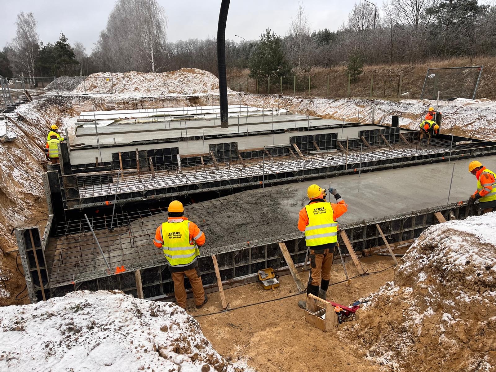 Wylewanie betonu na zbrojenie fundamentów budynku przez ekipę budowlaną w żółtych kamizelkach odblaskowych, widoczne szalunki i rura do podawania betonu, zimowa sceneria z resztkami śniegu.