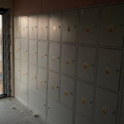 ,,BRODA-BUD” PRZEMYSŁAW MASTERNAK - Szereg szarych skrzynek elektrycznych z ostrzeżeniami, zamontowanych na ścianie w surowym stanie, widoczne płyty kartonowo-gipsowe na górze ściany, resztki materiałów budowlanych na podłodze.