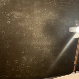 ,,BRODA-BUD” PRZEMYSŁAW MASTERNAK - Ściana pokryta dekoracyjną, ciemną fakturą z widocznymi nierównościami i wzorami, oświetlona przez lampę studyjną. Podłoga zabezpieczona folią malarską, widoczny fragment łóżka.