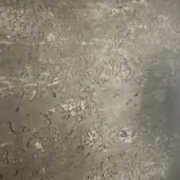 Szara ściana z nieregularną, chropowatą fakturą imitującą naturalny kamień lub beton, z widocznymi smugami i wgłębieniami.