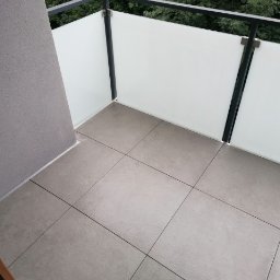 ,,BRODA-BUD” PRZEMYSŁAW MASTERNAK - Wykończony balkon z szarymi płytkami podłogowymi i białą szklaną balustradą z czarnymi elementami konstrukcyjnymi, widok z góry.