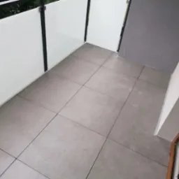 Wykończony balkon z szarymi płytkami podłogowymi, białą balustradą z czarnymi elementami i szarą ścianą w rogu.