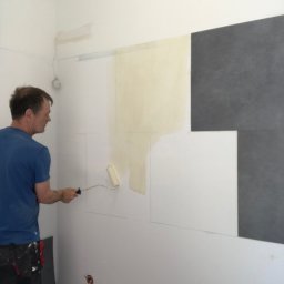 ,,BRODA-BUD” PRZEMYSŁAW MASTERNAK - Mężczyzna maluje ścianę wałkiem w pomieszczeniu z częściowo położonymi szarymi płytkami i zaznaczonymi liniami na ścianie, widoczna puszka z farbą i gniazdko elektryczne.
