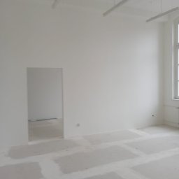 ,,BRODA-BUD” PRZEMYSŁAW MASTERNAK - Pusty, pomalowany na biało pokój biurowy w trakcie przygotowań do wykończenia, z widocznymi elementami podłogi zabezpieczonymi przed zabrudzeniem, oświetleniem sufitowym i oknem z grzejnikiem.