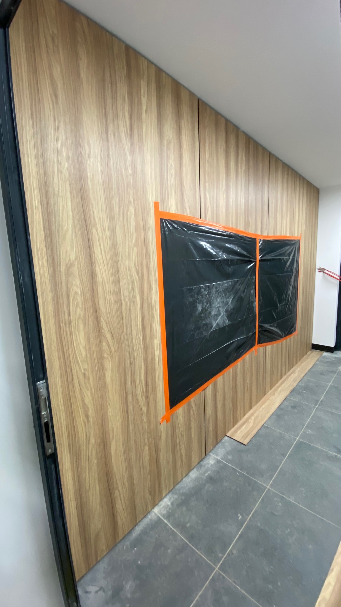 Wnętrze biura w trakcie prac wykończeniowych: ściana wyłożona panelami imitującymi drewno, zabezpieczone folią okna, szare płytki podłogowe.