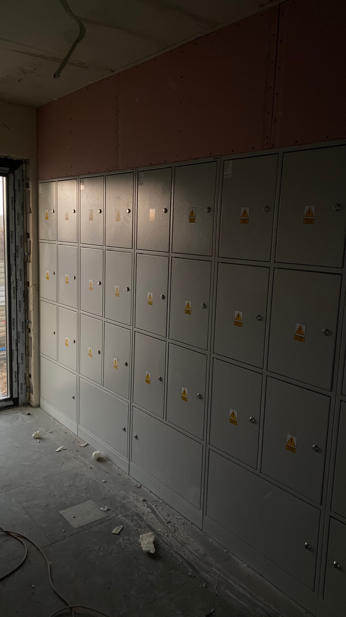 Szereg szarych skrzynek elektrycznych z ostrzeżeniami, zamontowanych na ścianie w surowym stanie, widoczne płyty kartonowo-gipsowe na górze ściany, resztki materiałów budowlanych na podłodze.