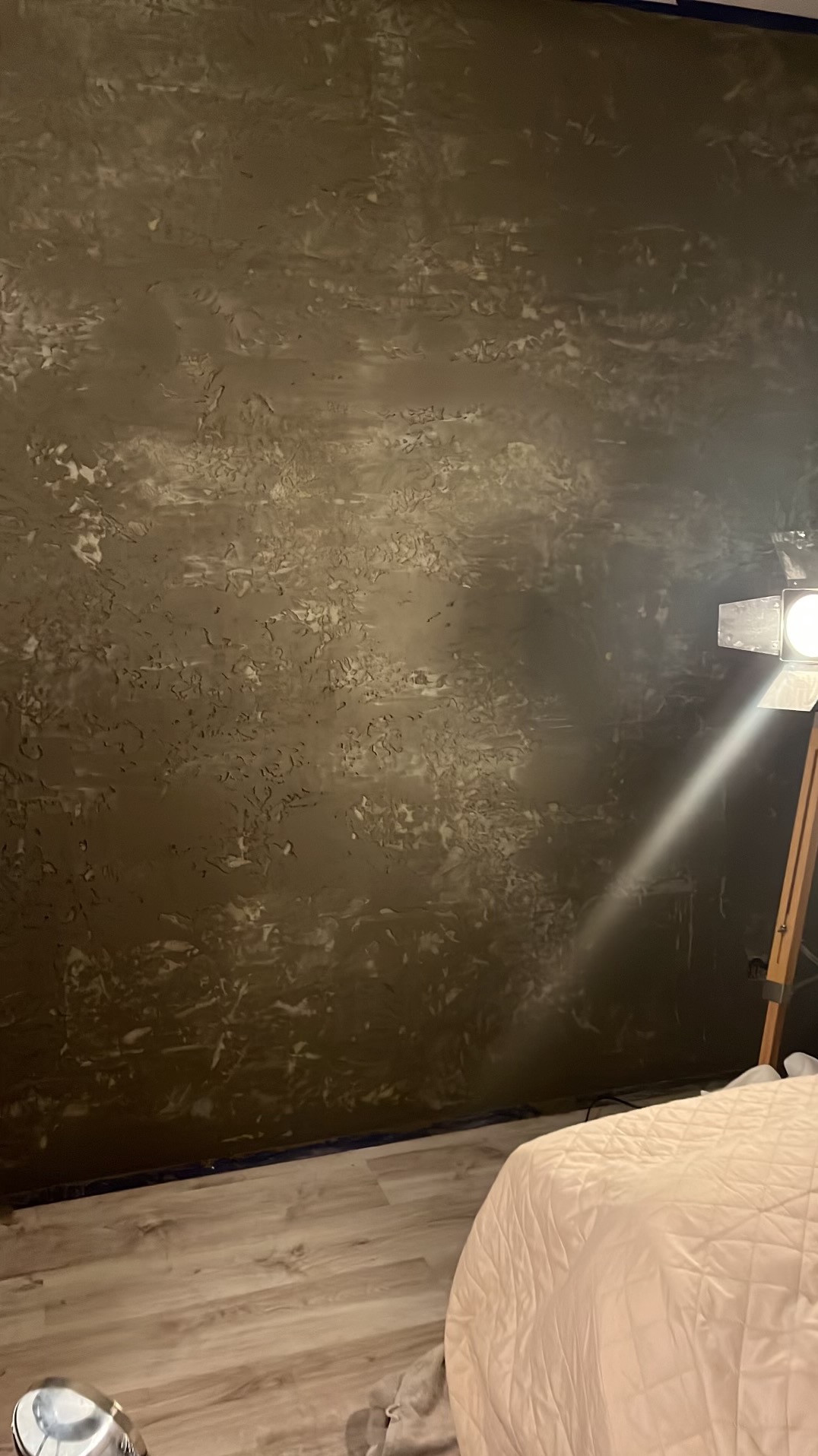 Ściana pokryta dekoracyjną, ciemną fakturą z widocznymi nierównościami i wzorami, oświetlona przez lampę studyjną. Podłoga zabezpieczona folią malarską, widoczny fragment łóżka.
