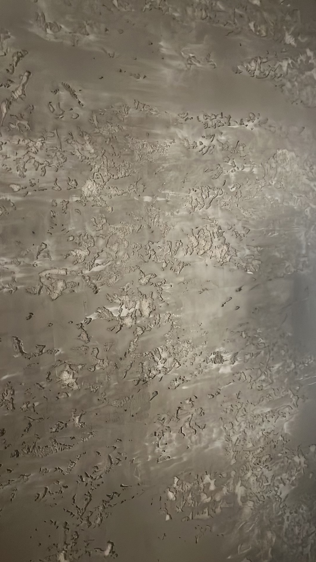 Szara ściana z nieregularną, chropowatą fakturą imitującą naturalny kamień lub beton, z widocznymi smugami i wgłębieniami.