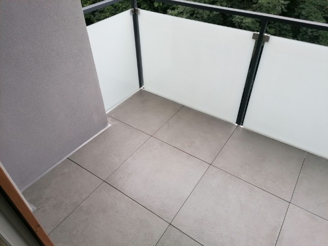 Wykończony balkon z szarymi płytkami podłogowymi i białą szklaną balustradą z czarnymi elementami konstrukcyjnymi, widok z góry.