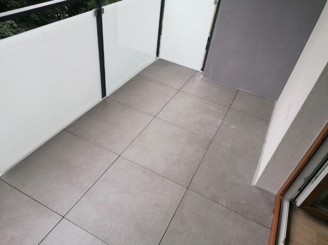 Wykończony balkon z szarymi płytkami podłogowymi, białą balustradą z czarnymi elementami i szarą ścianą w rogu.