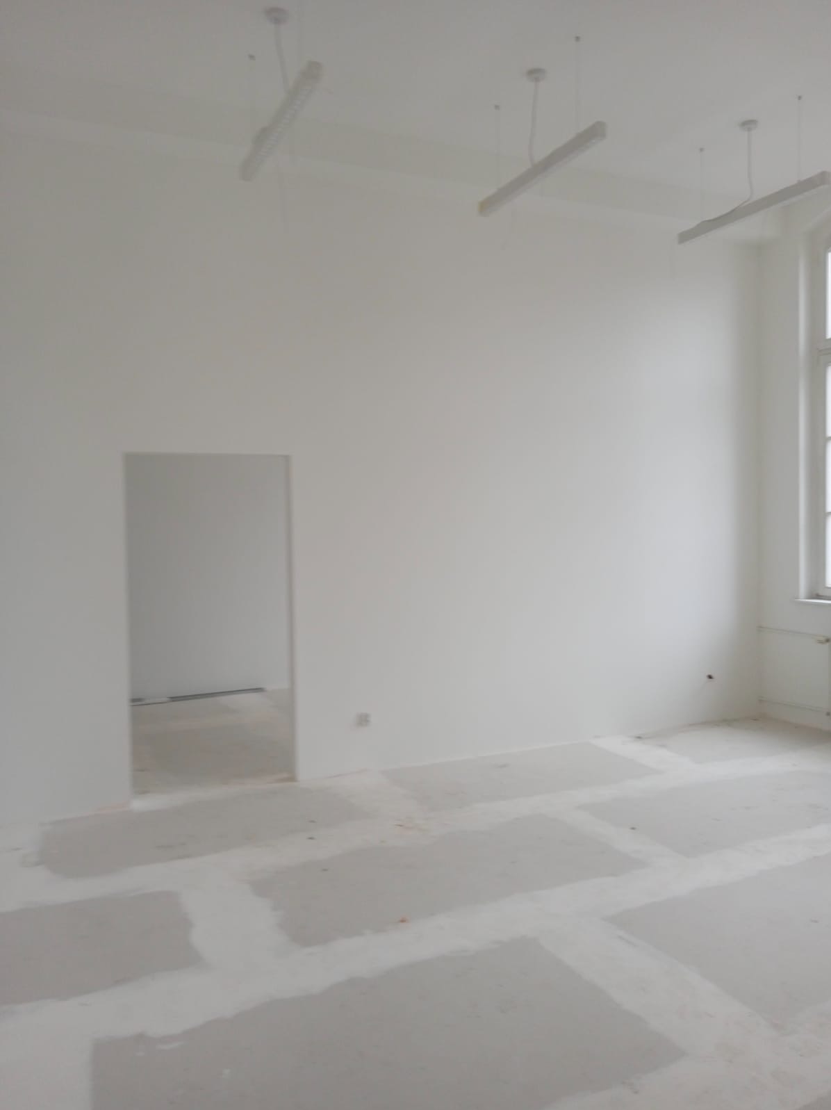 Pusty, pomalowany na biało pokój biurowy w trakcie przygotowań do wykończenia, z widocznymi elementami podłogi zabezpieczonymi przed zabrudzeniem, oświetleniem sufitowym i oknem z grzejnikiem.