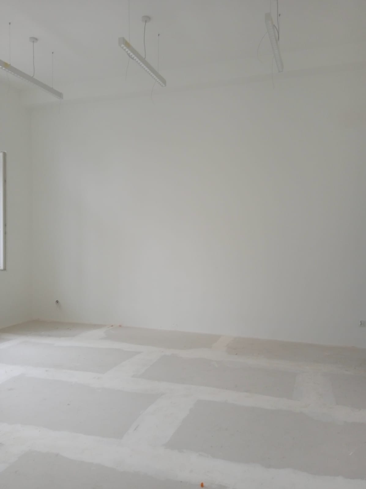 Wnętrze biura w trakcie remontu, widoczne gładkie, białe ściany, surowa podłoga z widocznymi śladami po gładzi, oraz wiszące lampy sufitowe.
