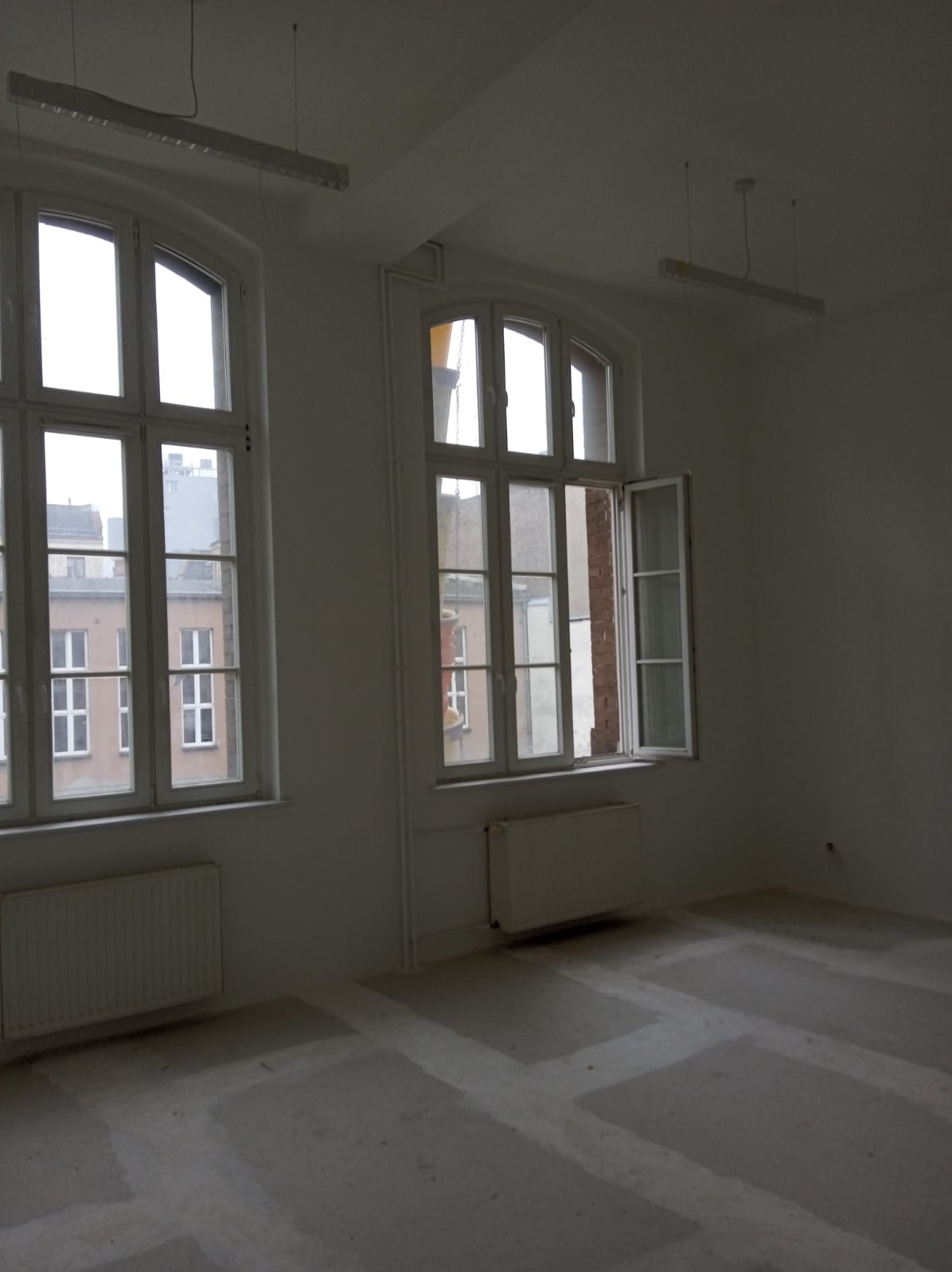 Puste, jasne pomieszczenie z dwoma dużymi oknami, grzejnikami i przygotowanym podłożem pod podłogę, widoczne elementy instalacji elektrycznej na suficie.