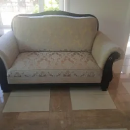 Dwuosobowa sofa tapicerowana w kremowo-złote wzory, z ciemnobrązowymi, lakierowanymi podłokietnikami i ramą, stojąca na jasnych, kwadratowych płytkach podłogowych.