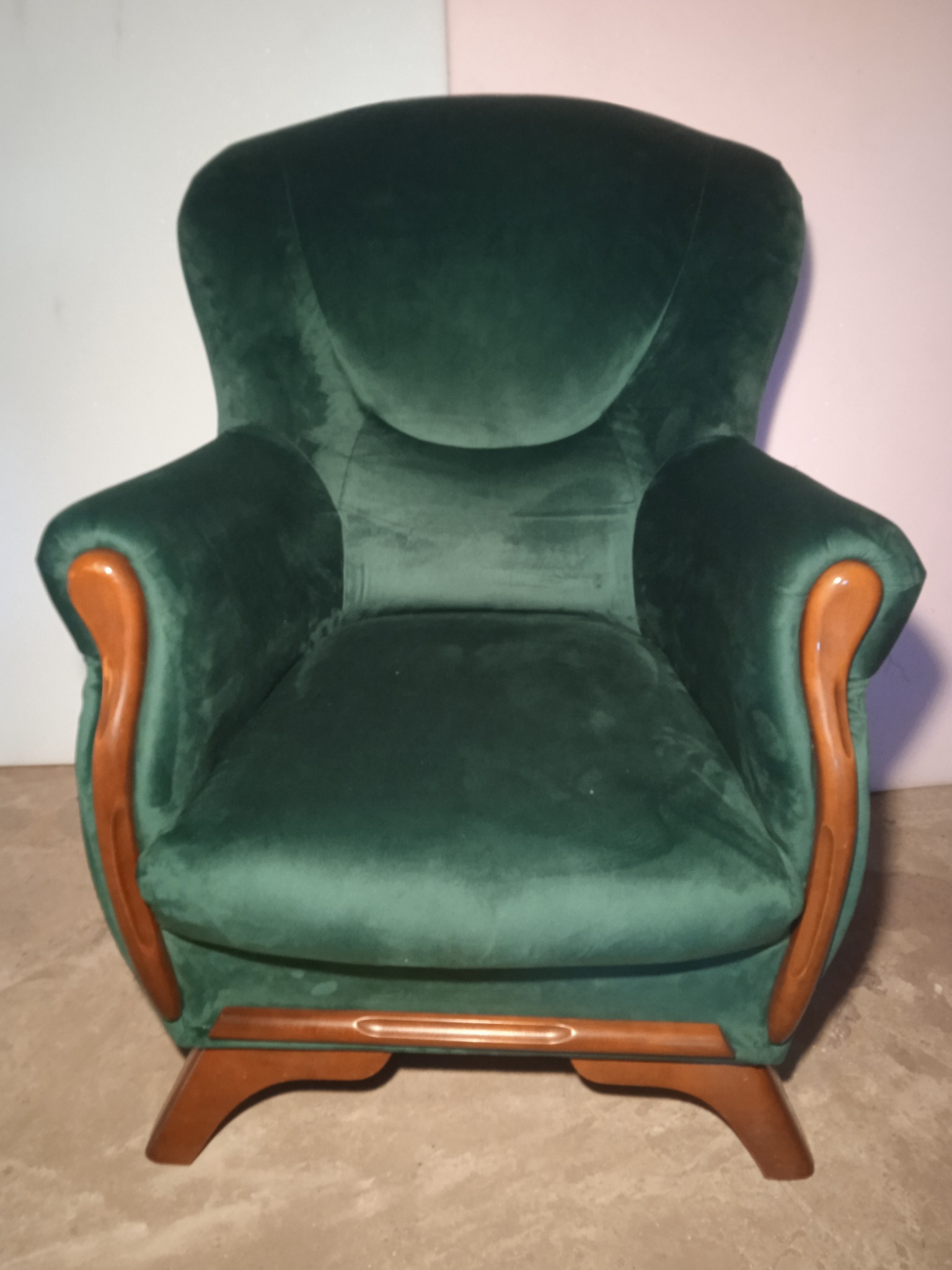 Zielony, welurowy fotel z drewnianymi podłokietnikami w stylu retro, ustawiony na szarej podłodze, z widocznym oświetleniem studyjnym.