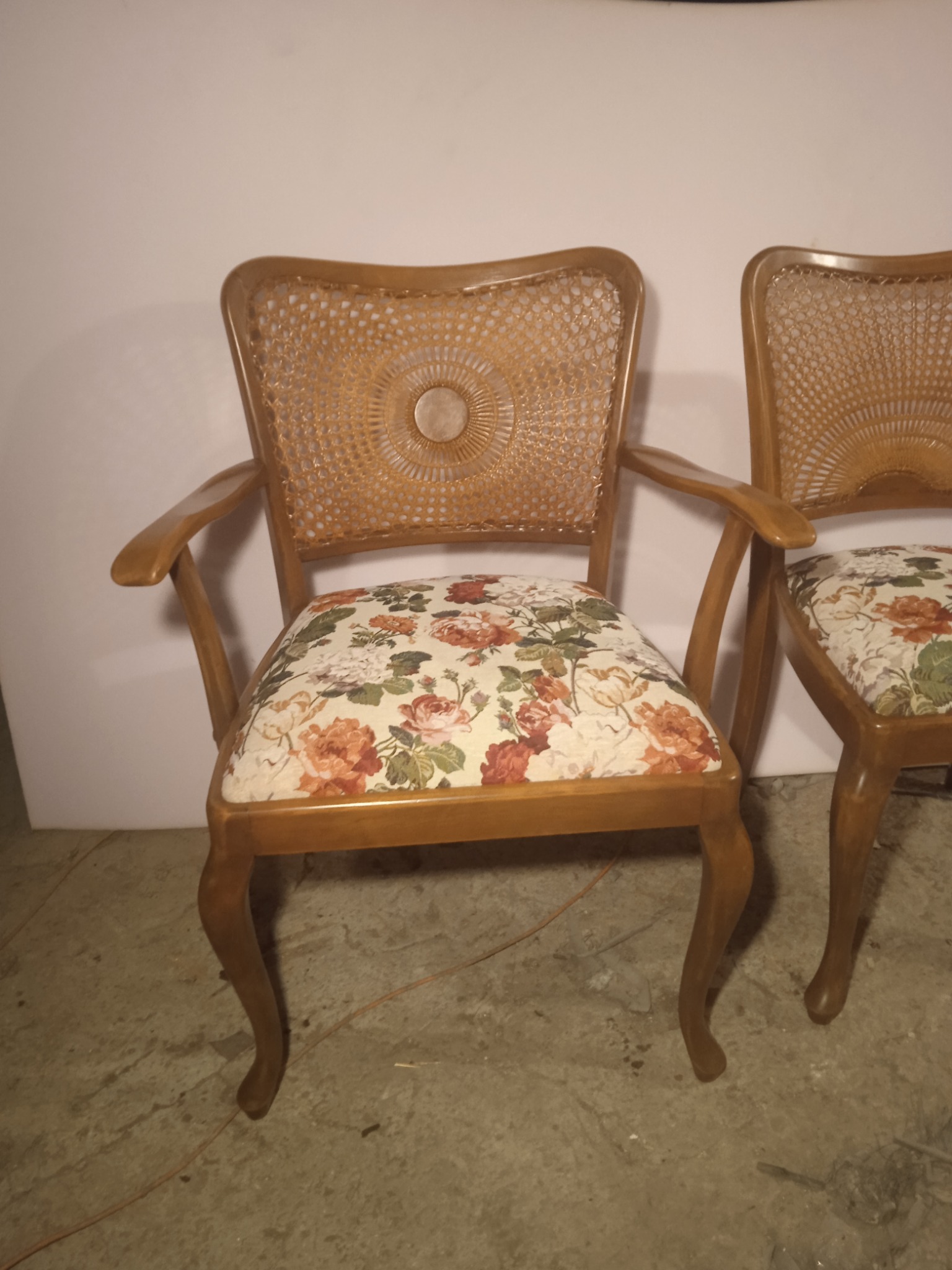 Dwa drewniane krzesła z plecionymi oparciami i tapicerowanymi siedziskami w kwiatowy wzór, ustawione na betonowej podłodze przed białą ścianą.