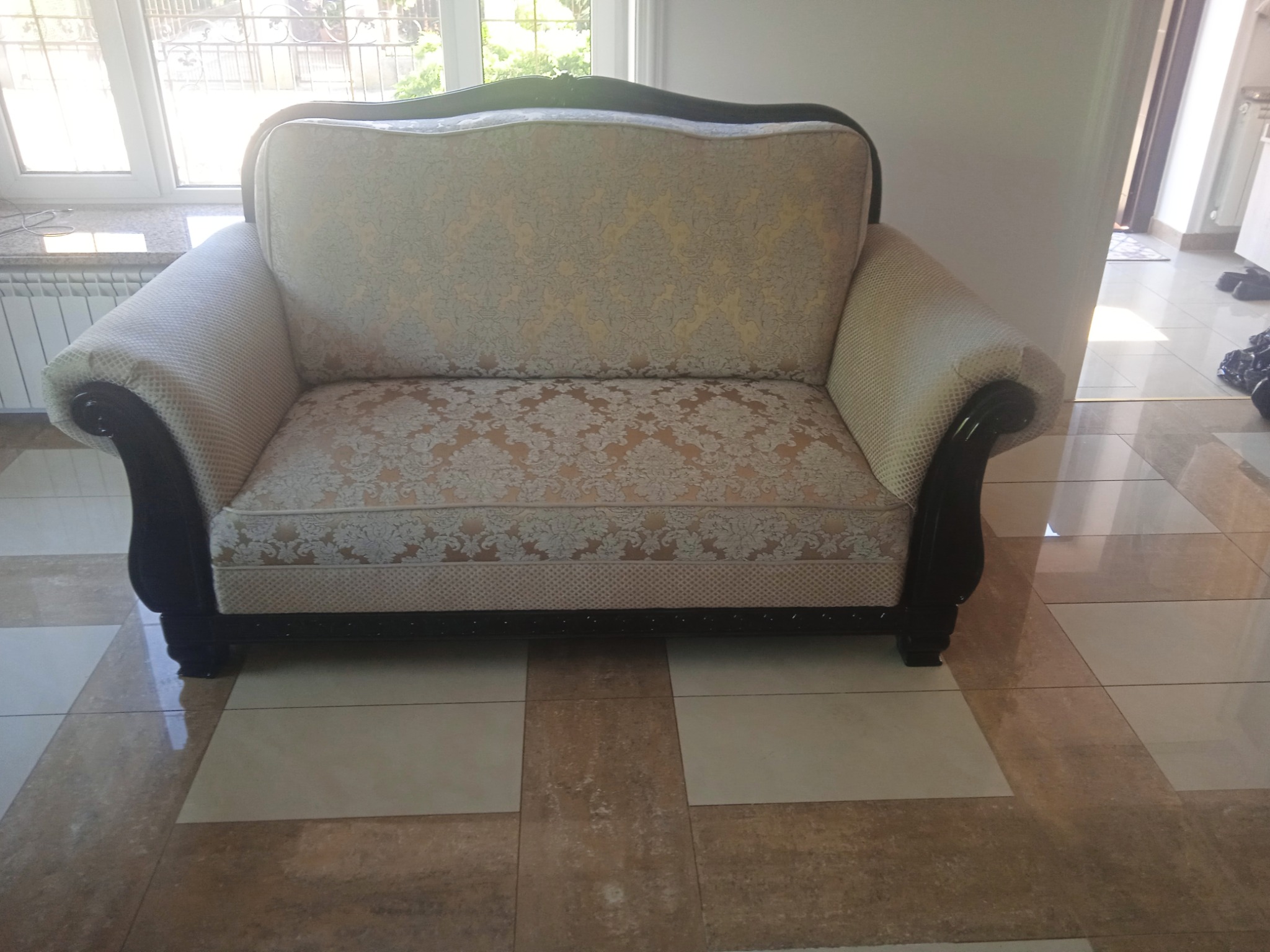 Dwuosobowa sofa tapicerowana w kremowo-złote wzory, z ciemnobrązowymi, lakierowanymi podłokietnikami i ramą, stojąca na jasnych, kwadratowych płytkach podłogowych.