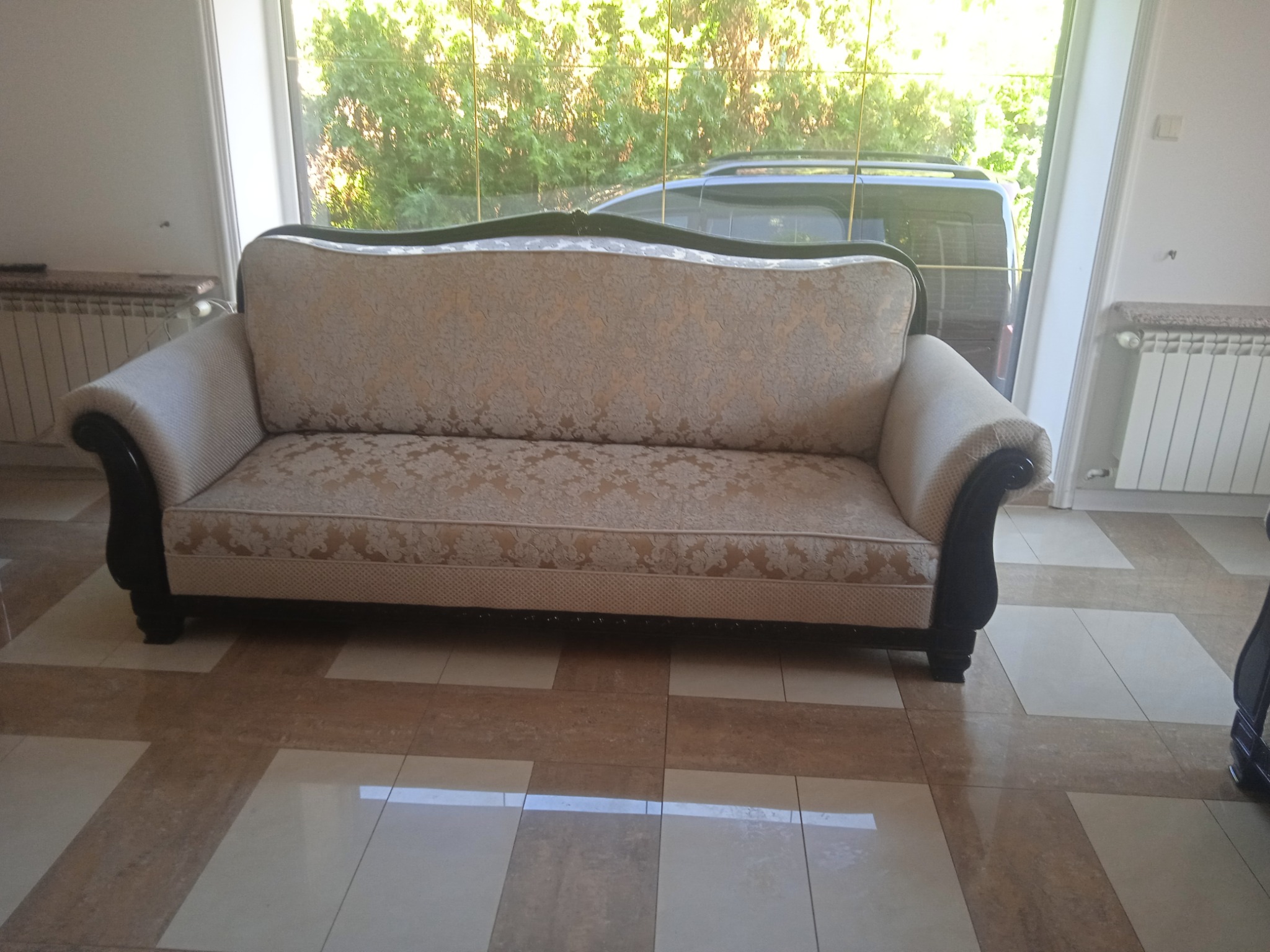 Dwuosobowa sofa z kremową tapicerką w kwiatowy wzór, z ciemnymi, rzeźbionymi bokami, ustawiona na błyszczącej podłodze z płytek w odcieniach beżu i brązu, z widokiem na zieleń za oknem.
