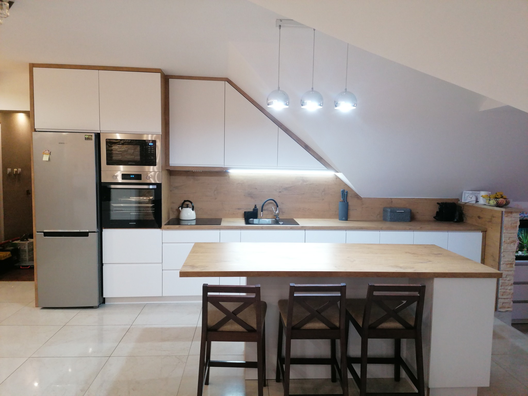 Nowoczesna kuchnia na poddaszu z białymi szafkami, drewnianym blatem, wyspą z trzema krzesłami i oświetleniem punktowym.