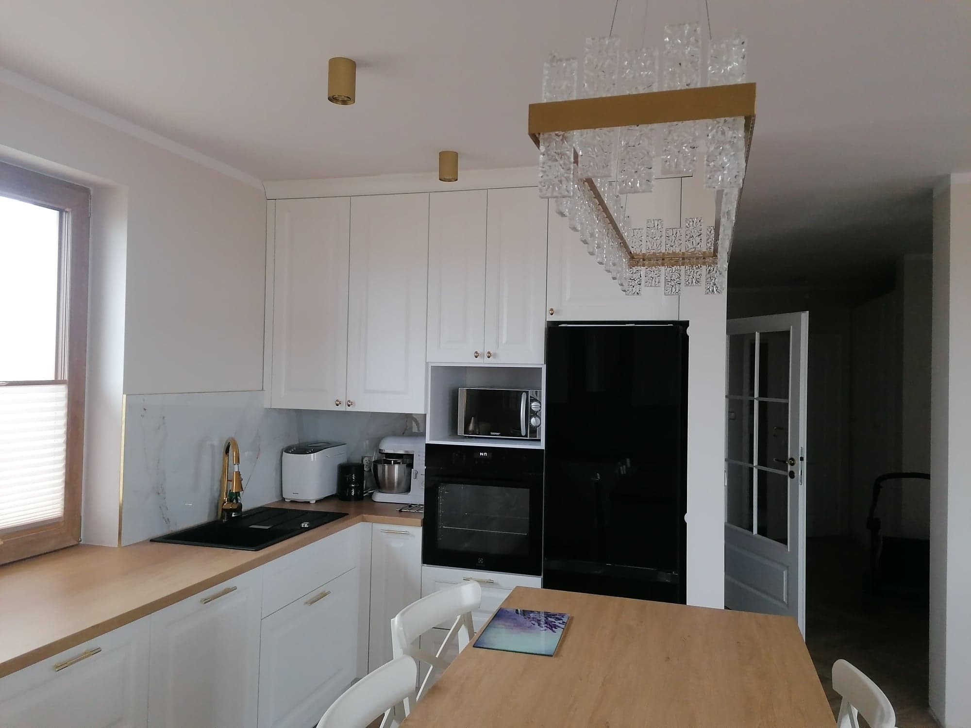 Jasna kuchnia z białymi szafkami, złotymi akcentami i kryształowym żyrandolem, widok na otwarte drzwi do innego pomieszczenia.