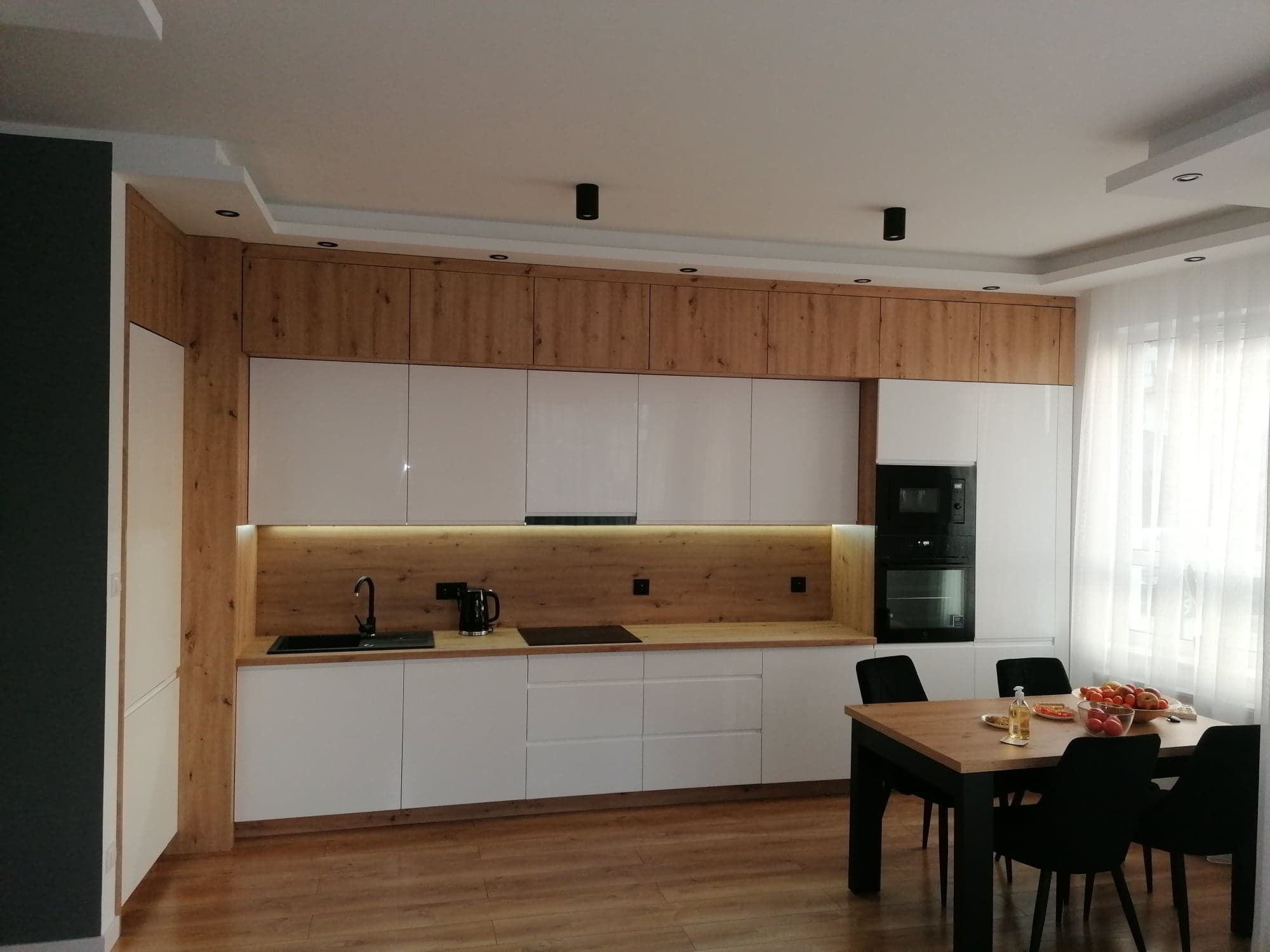 Nowoczesna kuchnia z białymi, lakierowanymi frontami i drewnianymi akcentami, oświetleniem LED pod szafkami górnymi i czarnymi elementami wykończeniowymi, widok na stół jadalny z owocami.