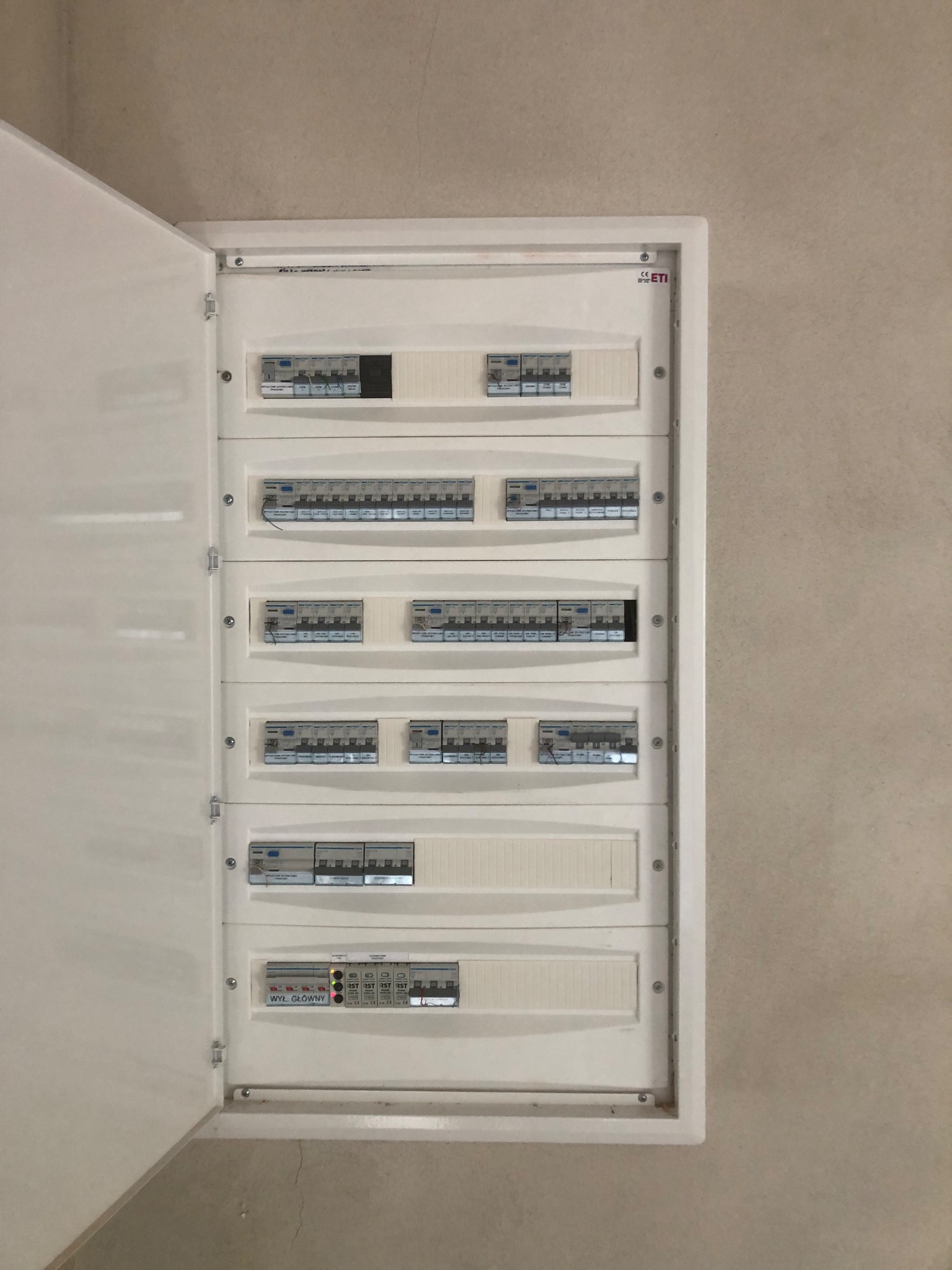 Otwarta, biała skrzynka elektryczna z bezpiecznikami na beżowej ścianie. Widoczne rzędy zabezpieczeń i oznaczenia. Skrzynka zamontowana równo, schludna instalacja.