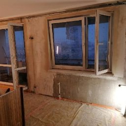IDEA HOUSE Przemysław Jurzec - Wnętrze w trakcie prac: widoczne okna balkonowe z otwartym skrzydłem, ściany w stanie surowym, puszki z farbą i przenośna lampa budowlana oświetlająca fragment podłogi.