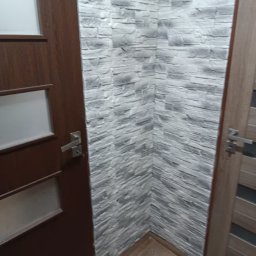 IDEA HOUSE Przemysław Jurzec - Fragment łazienki z wykończoną ścianą imitującą szary kamień oraz dwiema drewnianymi ościeżnicami drzwi po bokach.