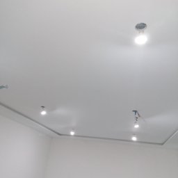 IDEA HOUSE Przemysław Jurzec - Biały sufit z zamontowanymi punktowymi lampami LED w Jastrzębiu-Zdroju, widoczne przewody przy jednym z otworów na oświetlenie i fragment drabiny w rogu kadru.