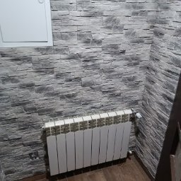 IDEA HOUSE Przemysław Jurzec - Ściana wykończona szarą okładziną imitującą kamień, z białym grzejnikiem panelowym na tle drewnianej podłogi i białej klapy rewizyjnej w górnym rogu.