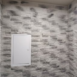 IDEA HOUSE Przemysław Jurzec - Ściana wyłożona szarymi panelami imitującymi kamień z ukrytymi drzwiczkami rewizyjnymi, widoczne nierówności i cieniowanie paneli.