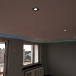 IDEA HOUSE Przemysław Jurzec - Pokój po remoncie z podwieszonym sufitem, oświetleniem LED w kolorach niebieskim i zielonym oraz punktowymi lampami sufitowymi, widok na osiedle przez okno.