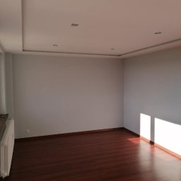 IDEA HOUSE Przemysław Jurzec - Pusty pokój z ciemną podłogą, świeżo pomalowanymi ścianami i grzejnikiem przy ścianie, wpadające promienie słoneczne przez okno.