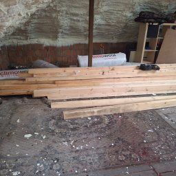 IDEA HOUSE Przemysław Jurzec - Poddasze z ocieploną pianką ścianą, ceglanym fragmentem i drewnianymi belkami leżącymi na dywanie przed regałem z narzędziami i materiałami budowlanymi.