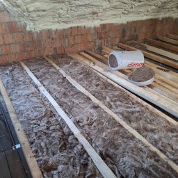 IDEA HOUSE Przemysław Jurzec - Ocieplenie poddasza wełną mineralną między legarami, widoczny fragment ściany z czerwonej cegły i puszka z wełną mineralną marki Climawool