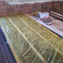 IDEA HOUSE Przemysław Jurzec - Izolacja z wełny mineralnej, zabezpieczona folią, ułożona między drewnianymi legarami na poddaszu, widoczny fragment muru z cegły, puszka z resztkami izolacji i narzędzie.