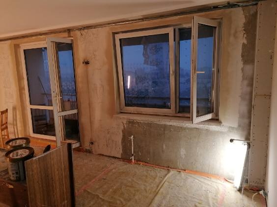 Wnętrze w trakcie prac: widoczne okna balkonowe z otwartym skrzydłem, ściany w stanie surowym, puszki z farbą i przenośna lampa budowlana oświetlająca fragment podłogi.