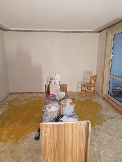 Pokój w trakcie prac przygotowawczych do malowania: zabezpieczona folią podłoga, białe ściany, widoczny worek z gładzią, pędzle, wałki i farby na stole, krzesło w tle.