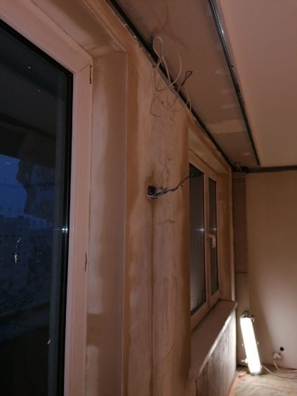 Wnętrze w trakcie przygotowań do malowania: ściany po nałożeniu gładzi gipsowej, widoczne przewody elektryczne, zamontowane profile sufitowe, okno z białymi ramami.