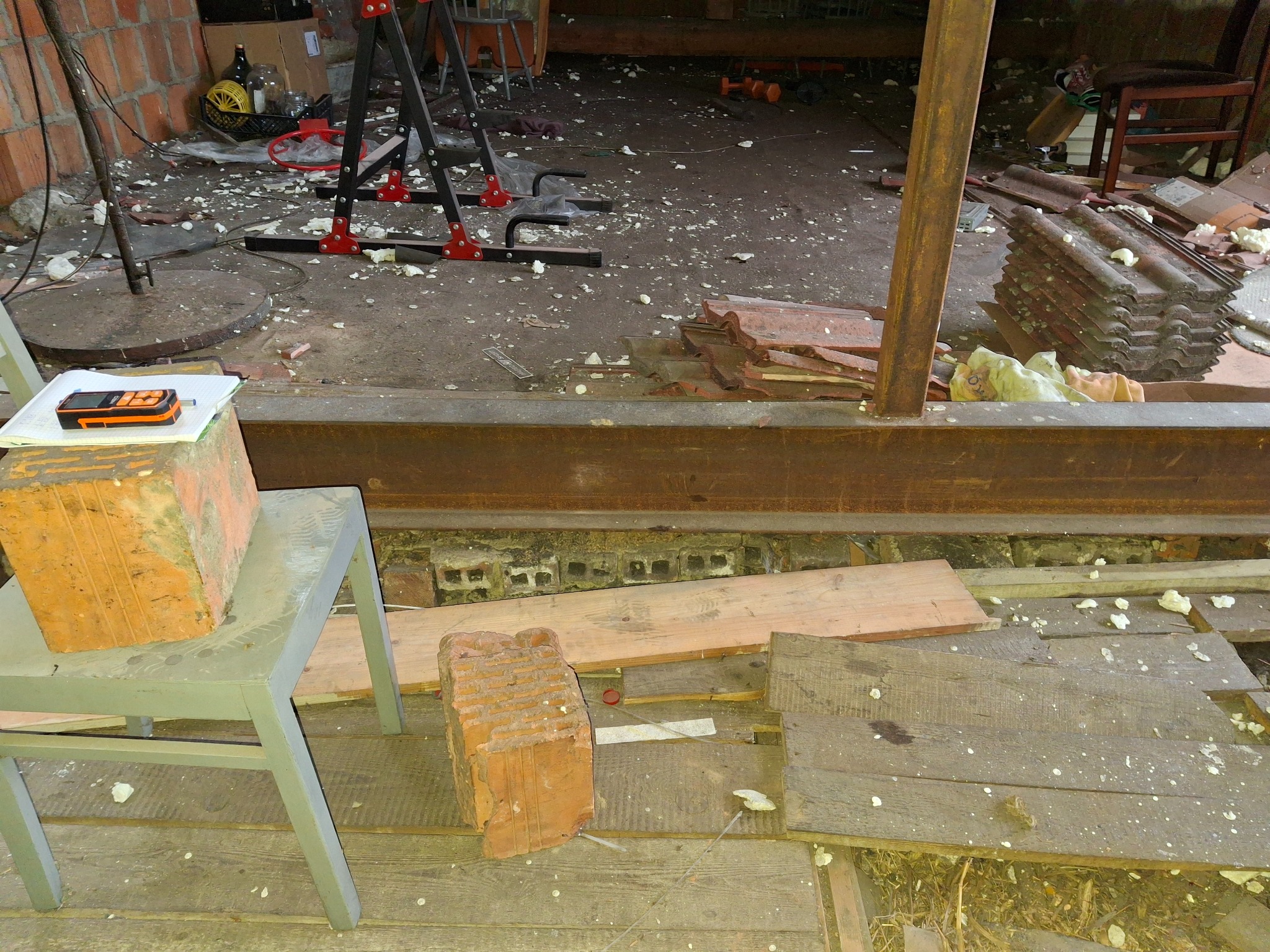 Wnętrze w trakcie prac remontowych: cegła na krześle z dalmierzem, widoczne belki stalowe, stare deski podłogowe i gruz z pianki montażowej.