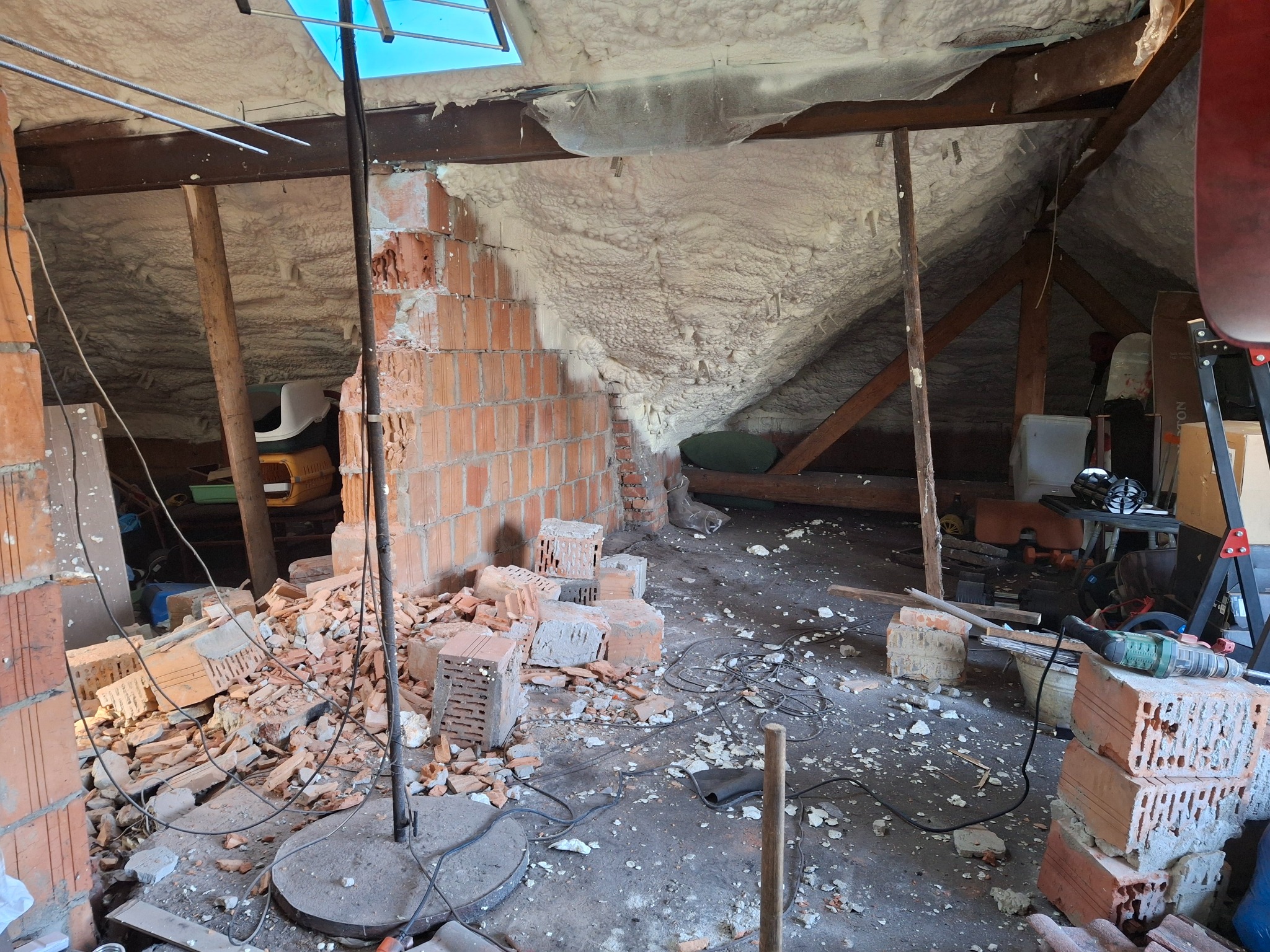 Poddasze w trakcie remontu: widoczna ceglana ściana w budowie, izolacja piankowa na dachu, porozrzucane cegły, narzędzia i kable na podłodze