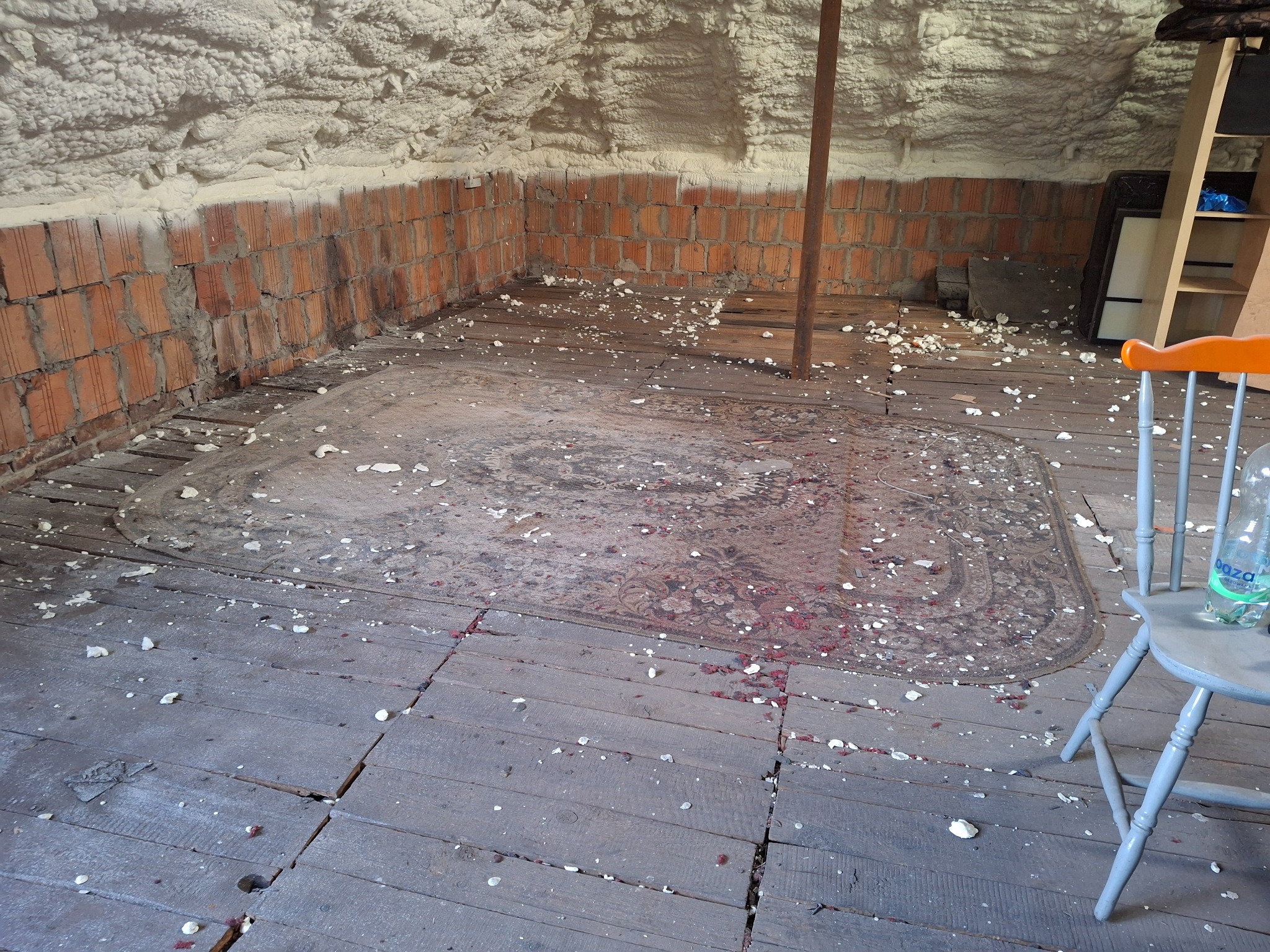 Poddasze z ocieplonym stropem pianką PUR, surową cegłą i drewnianą podłogą pokrytą starym dywanem, widoczne pozostałości pianki po aplikacji.