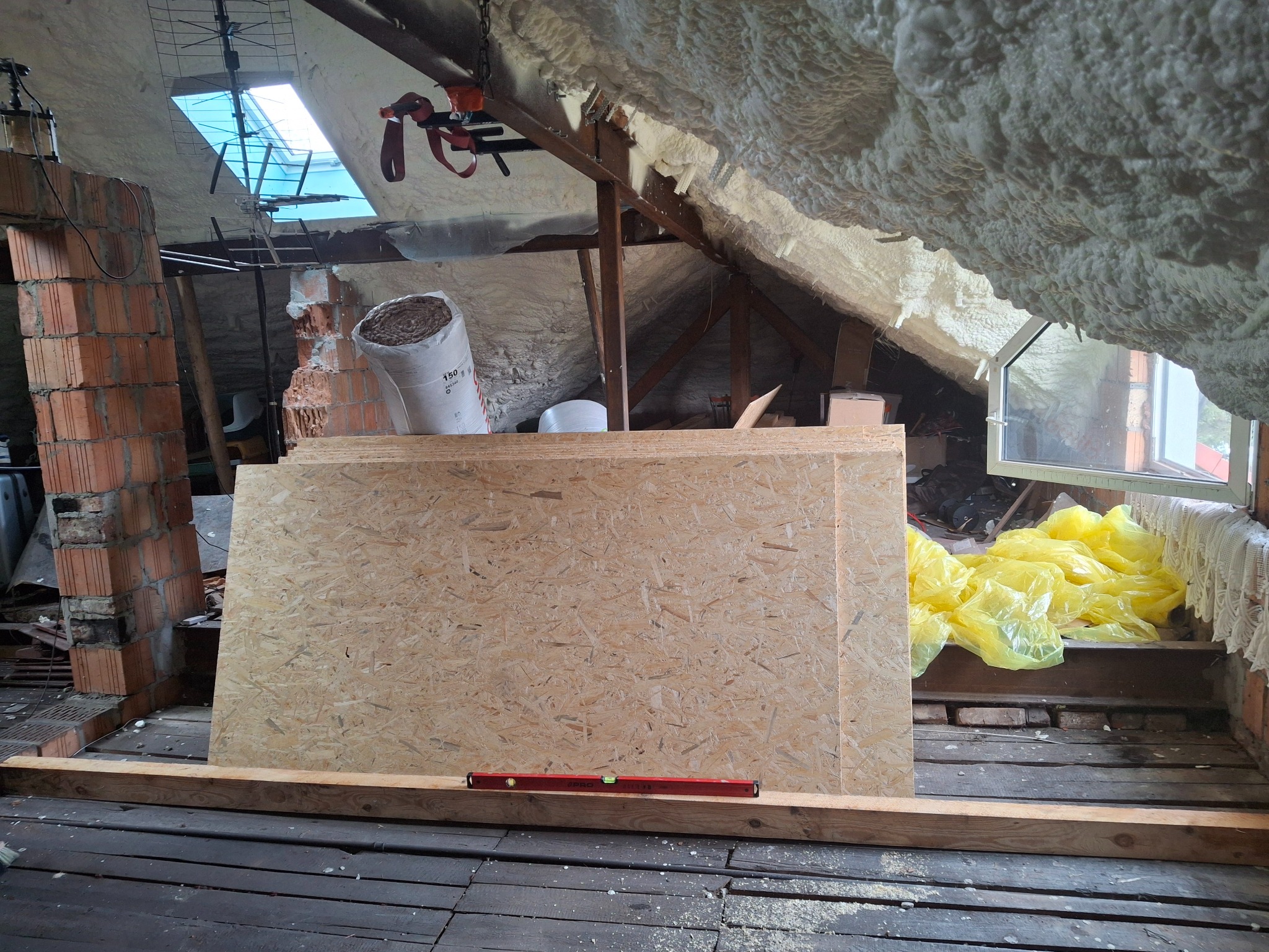 Płyty OSB o jasnej strukturze ułożone w stos na drewnianej belce w trakcie prac na poddaszu, widoczny poziomica, ceglana ściana w tle i ocieplenie pianką na dachu.