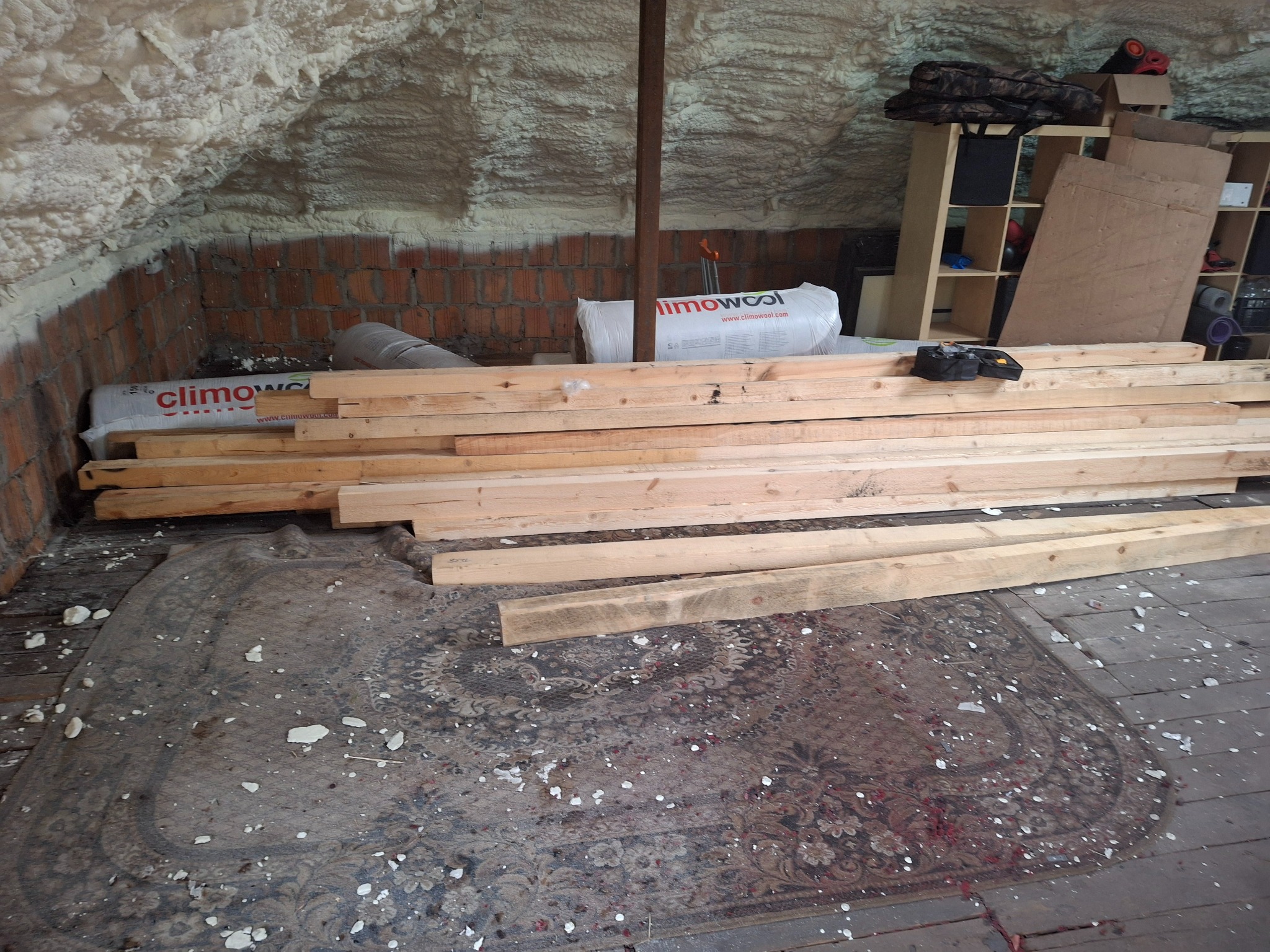 Poddasze z ocieploną pianką ścianą, ceglanym fragmentem i drewnianymi belkami leżącymi na dywanie przed regałem z narzędziami i materiałami budowlanymi.