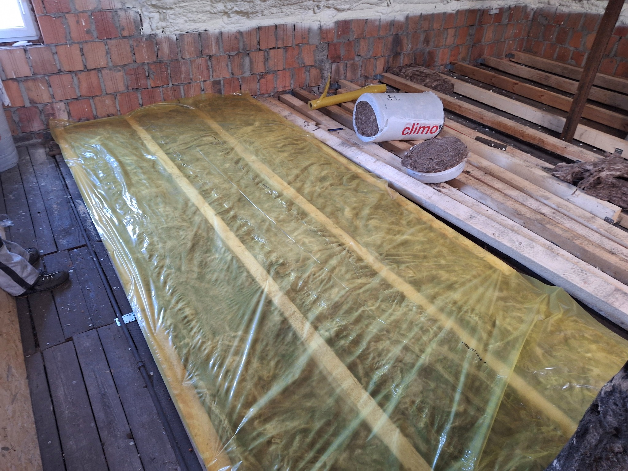 Izolacja z wełny mineralnej, zabezpieczona folią, ułożona między drewnianymi legarami na poddaszu, widoczny fragment muru z cegły, puszka z resztkami izolacji i narzędzie.