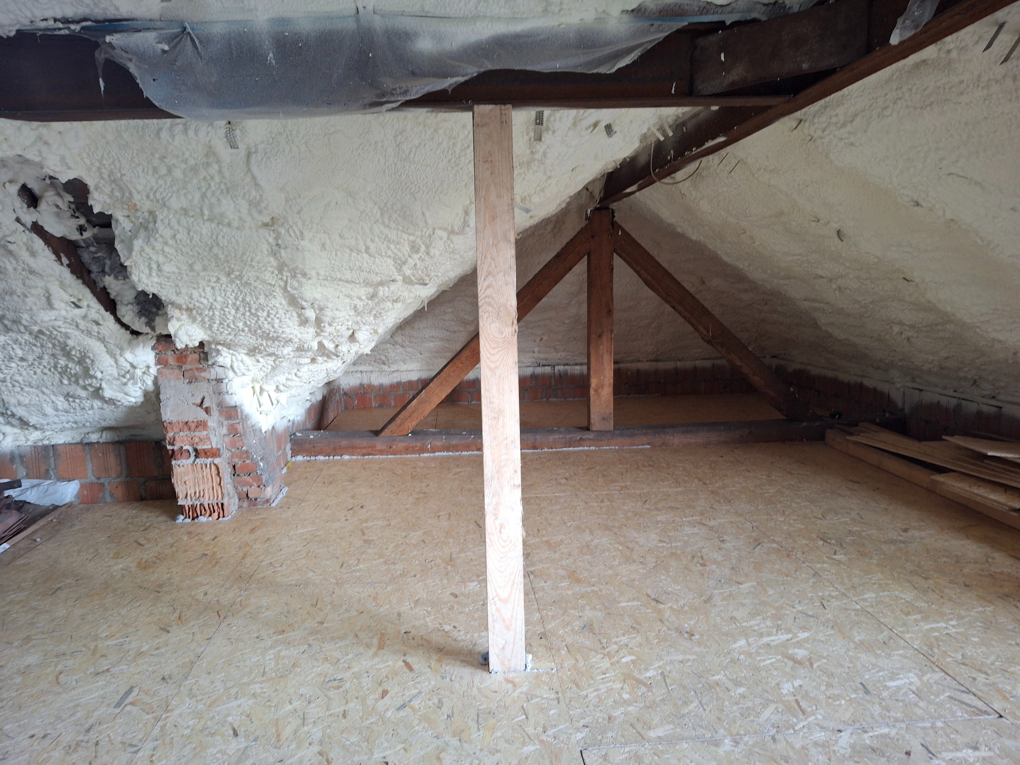 Poddasze w trakcie ocieplania pianą PUR, widoczne krokwie, mur z czerwonej cegły i drewniana podłoga z płyt OSB.