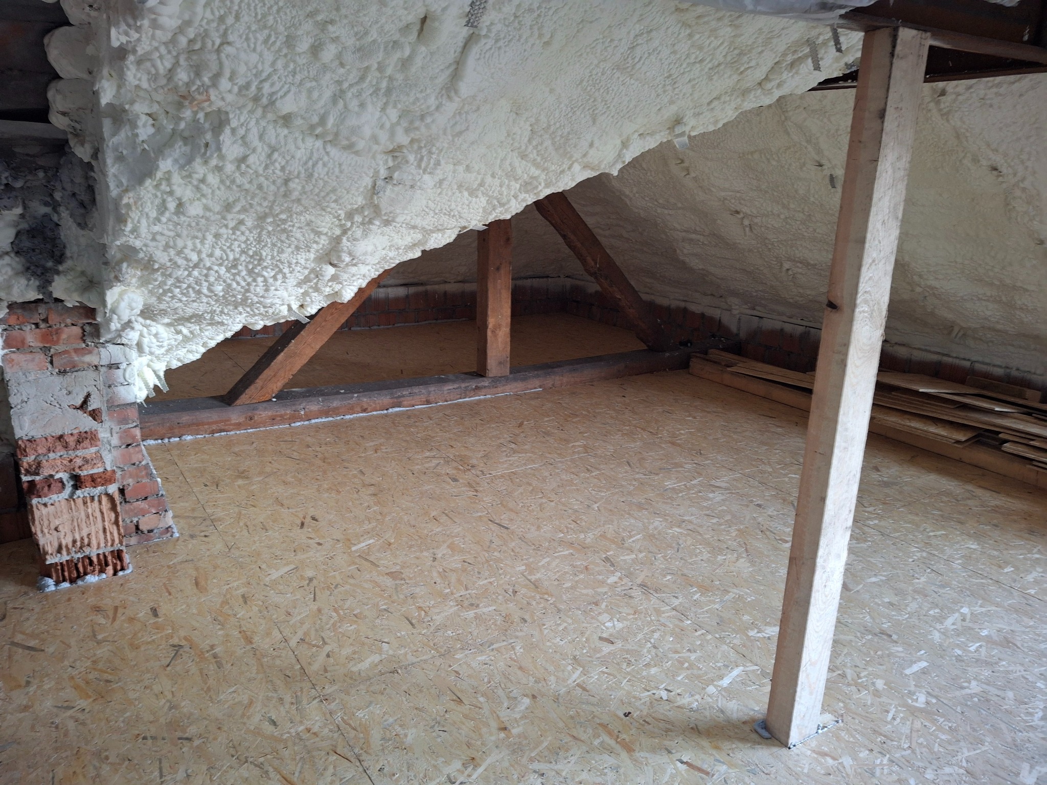 Poddasze ocieplone pianą PUR, widoczne elementy konstrukcyjne dachu i podłoga z płyt OSB, fragment ściany z cegły. Izolacja natryskowa na skośnej powierzchni.